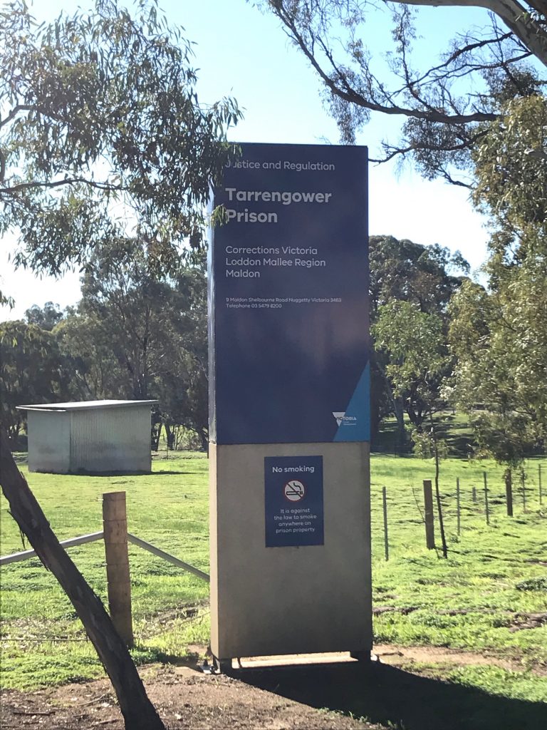 Sign at the entrance to Tarrengower Prison in Maldon, Victoria, operated by Corrections Victoria, with surrounding trees and open land.